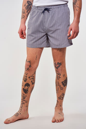 Pantalon de bain rayé bleu de Le Fabuleux Marcel de Bruxelles, avec des tatouages sur les jambes et une ceinture à la taille.