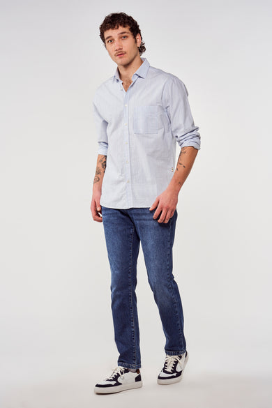 Chemise bleue à manches longues de Le Fabuleux Marcel de Bruxelles, portée avec des jeans bleu marine et des baskets blanches.