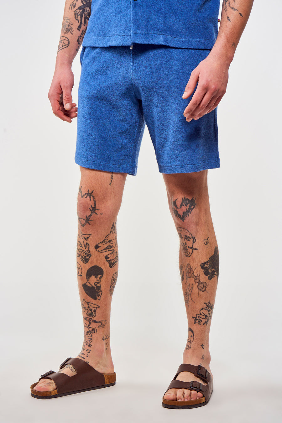 Short bleu de Le Fabuleux Marcel de Bruxelles, porté avec des sandales brunes et des tatouages visibles.