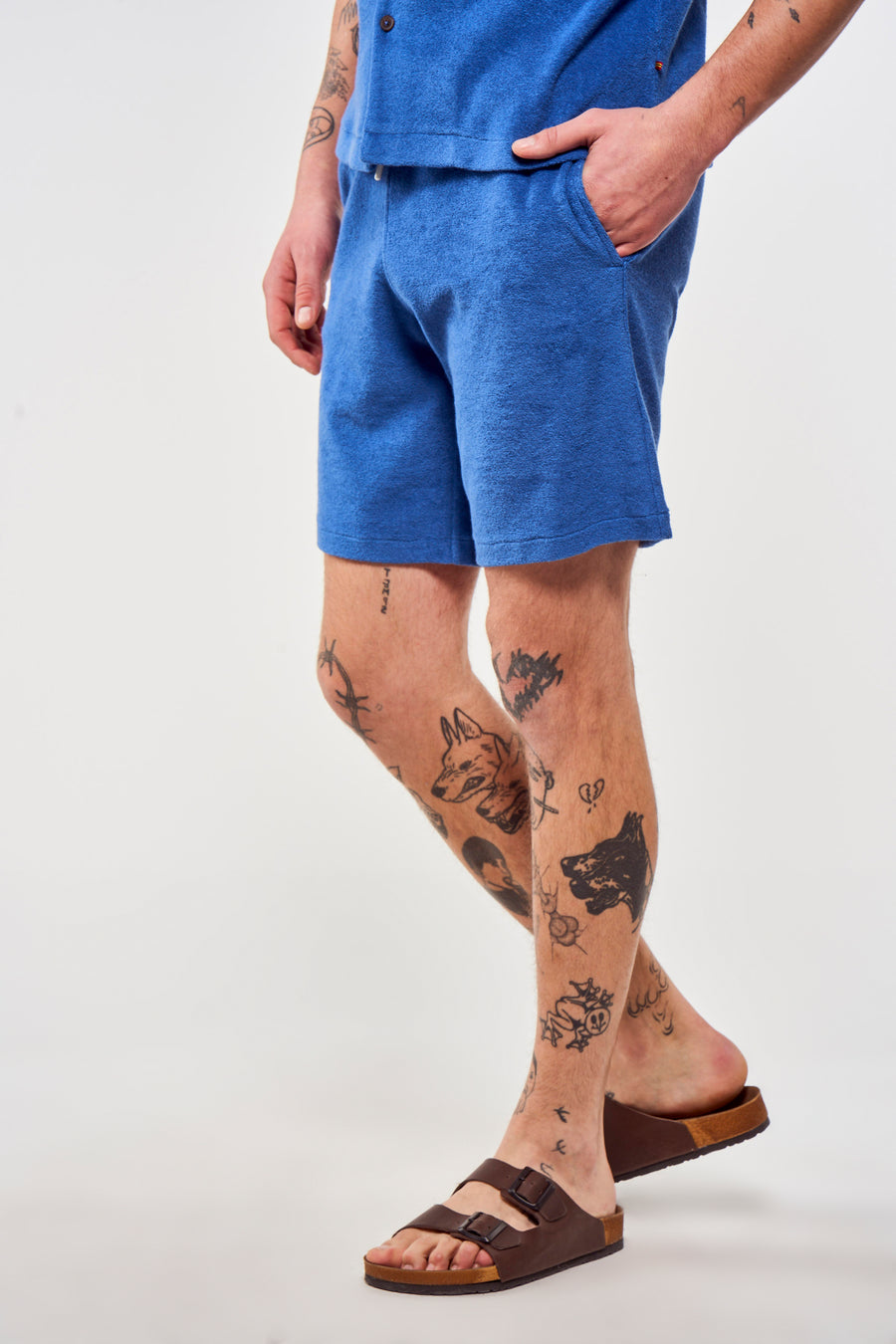 Blauwe short van Le Fabuleux Marcel de Bruxelles, gedragen met bruine sandalen en zichtbare tatoeages.
