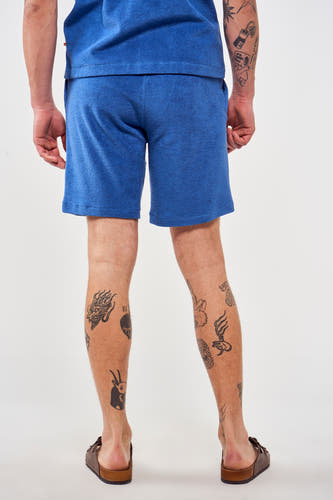 Short bleu de Le Fabuleux Marcel de Bruxelles, vu de derrière, avec des tatouages sur les jambes et des sandales brunes.