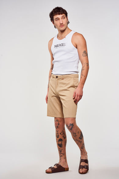 Short marron de Le Fabuleux Marcel de Bruxelles, associé à une chemise blanche et des sandales marrons, avec des tatouages sur les jambes.