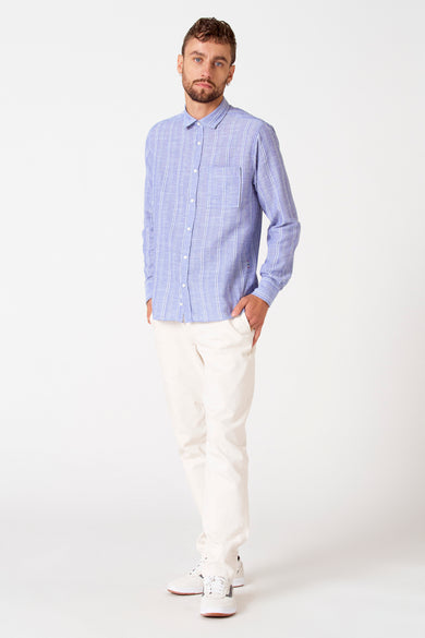 Chemise rayée bleue de longues manches de Le Fabuleux Marcel de Bruxelles, portée avec un pantalon crème et des baskets blanches.