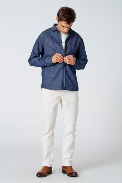 Chemise bleue à longues manches de Le Fabuleux Marcel de Bruxelles, avec un motif à carreaux subtil, portée avec un T-shirt blanc, des pantalons crème et des bottes brunes.