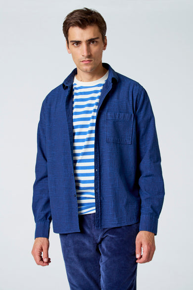 Chemise bleue à manches longues de Le Fabuleux Marcel de Bruxelles, portée sur un chemisier rayé.