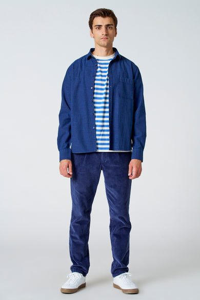 Bleu, chemise de lin à manches longues de Le Fabuleux Marcel de Bruxelles, portée sur une chemise rayée avec un pantalon en corduroy et des baskets blanches.