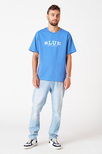T-shirt bleu à manches courtes de Le Fabuleux Marcel de Bruxelles, porté avec un jean léger et des baskets blanches.