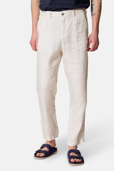 Pantalon beige Le Fabuleux Marcel de Bruxelles, avec des plis et des sandales bleues.
