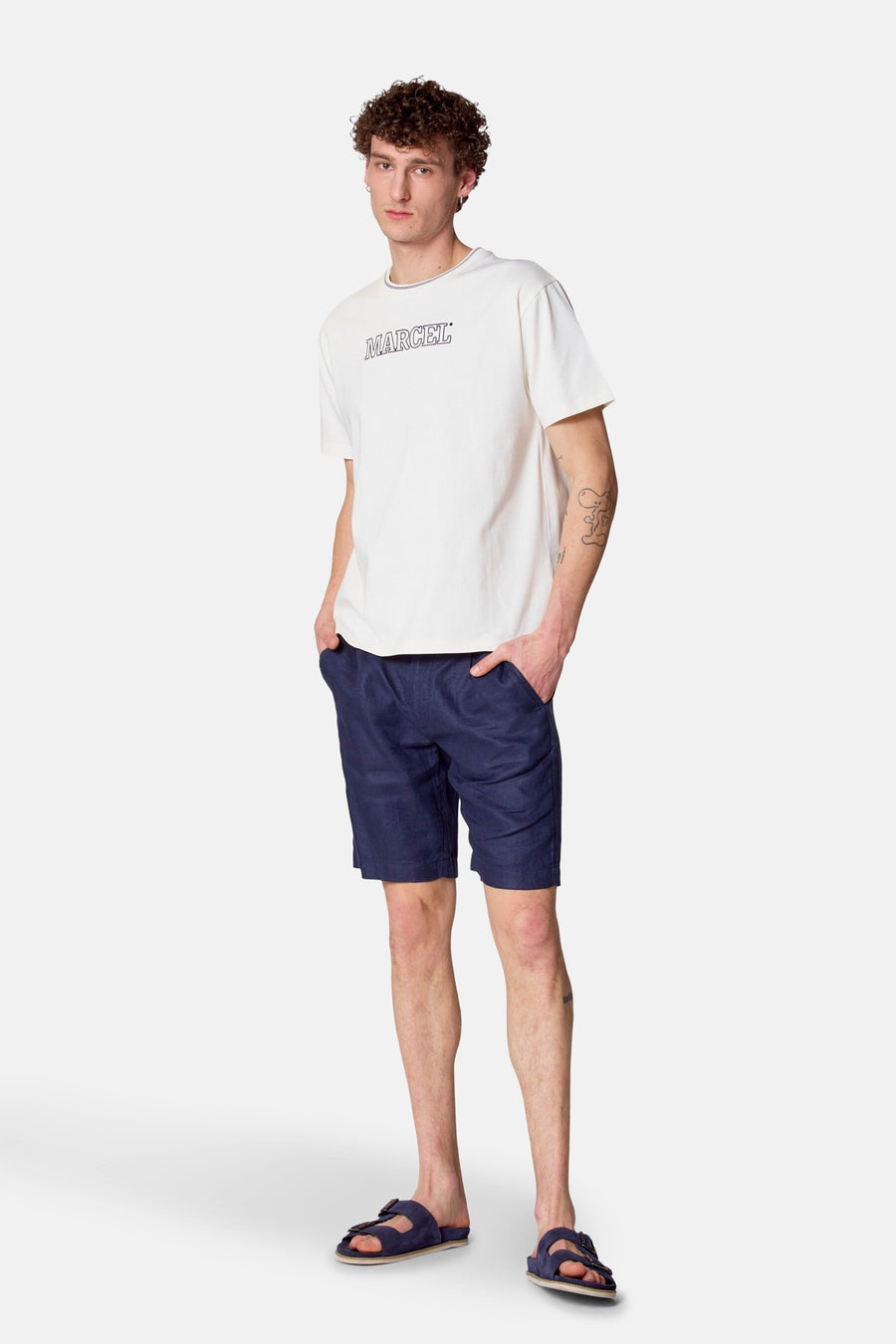 Man in witte T-shirt, blauwe shorts en sandalen, staand.