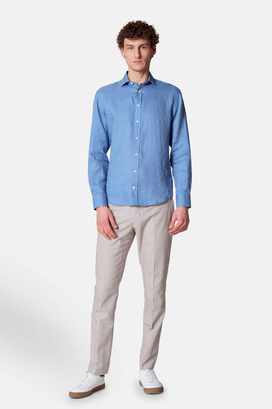 Man met blauw linnen overhemd en geruite broek, witte sneakers, staand.
