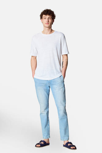 T-shirt blanc à manches courtes de Le Fabuleux Marcel de Bruxelles, porté avec des jeans légers et des sandales bleues.