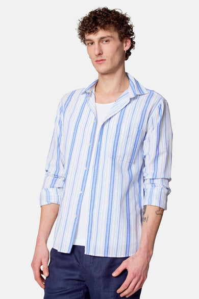 Chemise rayée bleue de Le Fabuleux Marcel de Bruxelles avec des manches longues, portée sur un T-shirt blanc et un pantalon bleu marine.