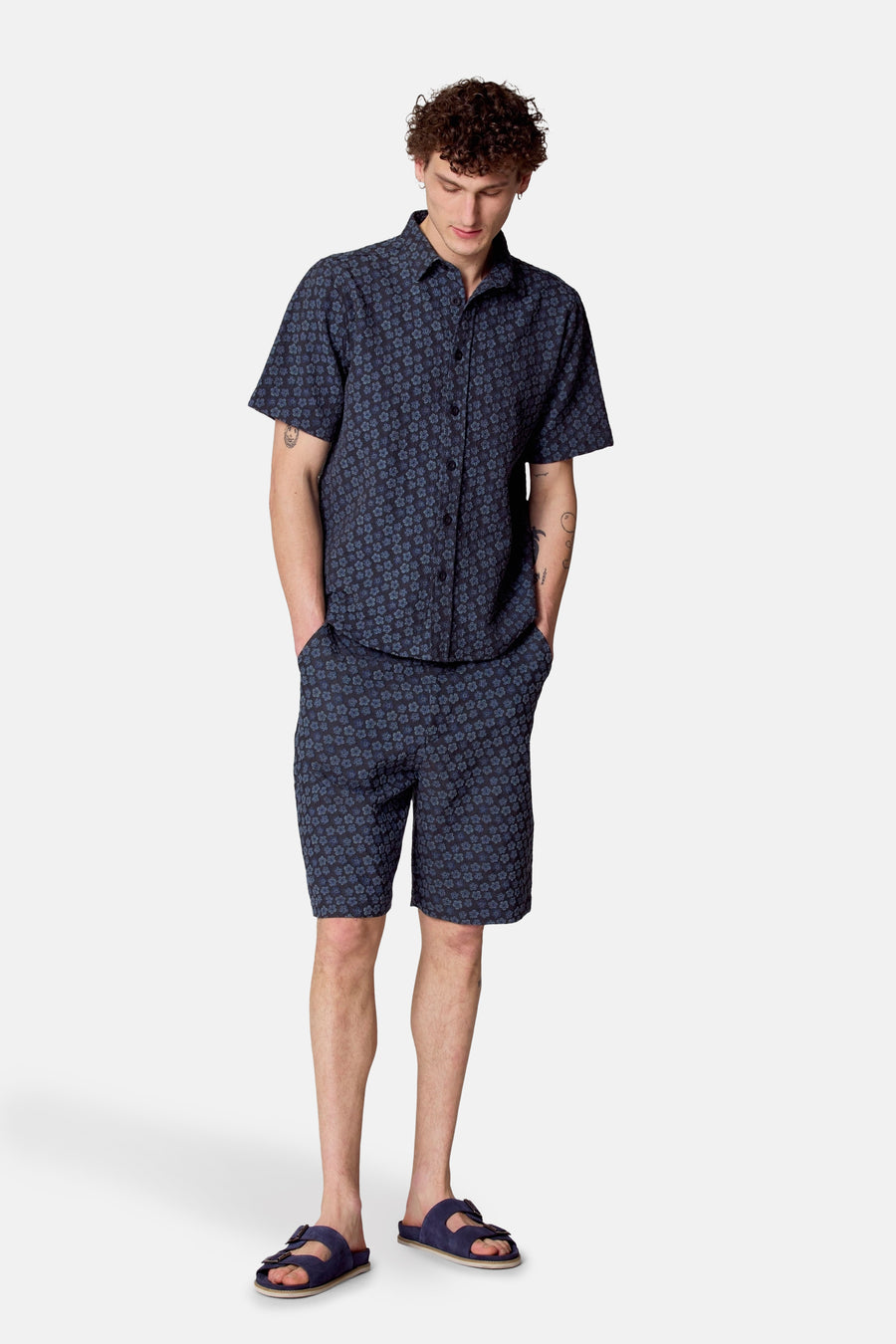 Een man in een bijpassende blauwe bloemenshirt en -short met blauwe sandalen, staand.