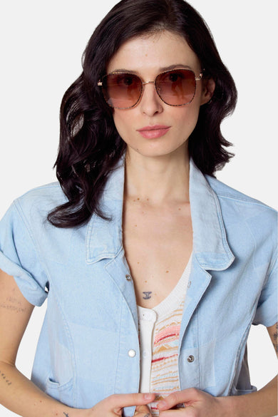 Lunettes de soleil roses de Le Fabuleux Marcel de Bruxelles, avec un cadre métallique et des verres marrons, portées par une femme en chemise bleu clair.