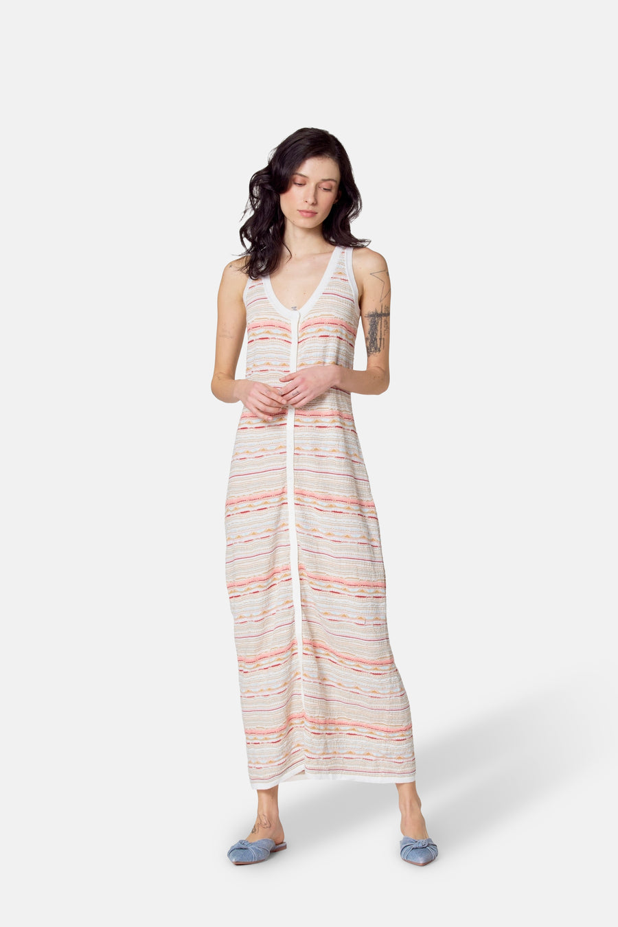 Vrouw met bruin haar in gestreepte jurk met knoopsluiting, blauwe muiltjes, witte achtergrond.