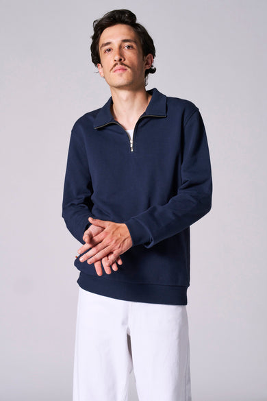 Pull bleu de Le Fabuleux Marcel de Bruxelles, avec une fermeture à glissière courte et porté avec un pantalon blanc.