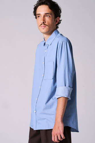 Chemise bleue à manches longues de Le Fabuleux Marcel de Bruxelles, avec des manches retroussées, portée avec un pantalon marron.