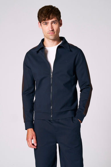 Veste bleue de Le Fabuleux Marcel de Bruxelles, avec une bande brune sur le poignet, portée sur un T-shirt blanc et un pantalon marine.