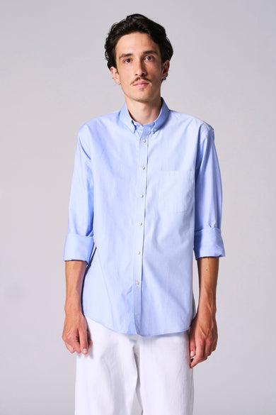Chemise bleue à manches longues de Le Fabuleux Marcel de Bruxelles, avec un gilet rayé et des manches retroussées, portée avec un pantalon blanc.