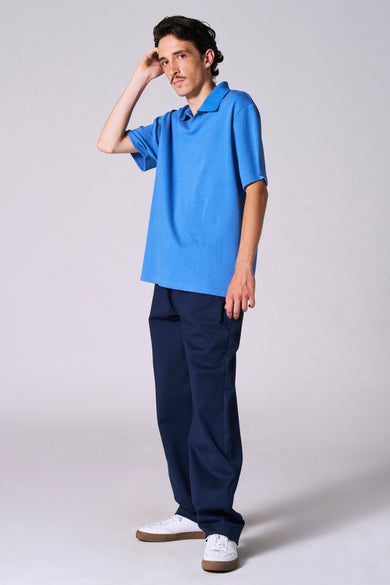 Polo bleu à manches courtes de Le Fabuleux Marcel de Bruxelles, porté avec un pantalon bleu marine et des baskets blanches.