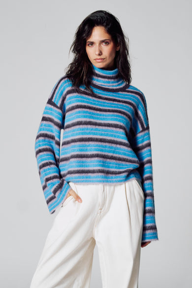 Jean à col montant bleu de Le Fabuleux Marcel de Bruxelles, avec un motif rayé et des manches longues, porté avec un pantalon blanc.