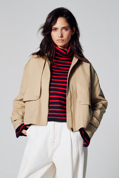 Manteau court beige de Le Fabuleux Marcel de Bruxelles, avec poches, associé à un pull rayé et des pantalons blancs.