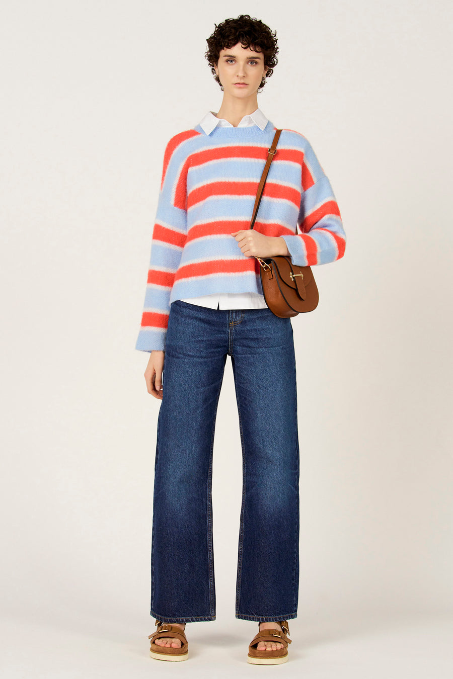 Pull multicolore à col rond de Le Fabuleux Marcel de Bruxelles, avec des rayures bleues et oranges sur un fond blanc, associé à un jean bleu foncé et un sac en cuir marron.