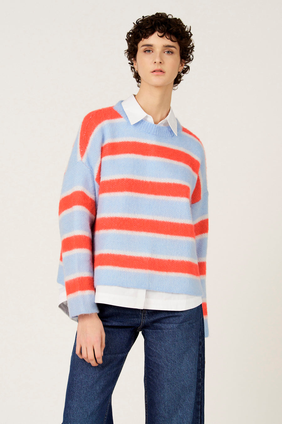Pull multicolore à col rond de Le Fabuleux Marcel de Bruxelles, motif rayé bleu et orange, porté sur une chemise blanche, avec un jean foncé.