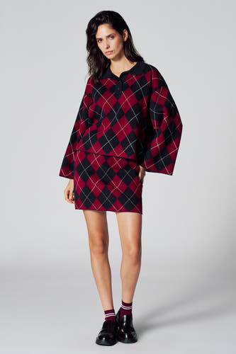 Short multicolor skirt from Le Fabuleux Marcel de Bruxelles, with a plaid pattern, combined with a sweater, socks, and black shoes.
