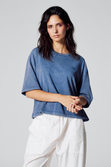 T-shirt bleu à manches courtes de Le Fabuleux Marcel de Bruxelles, porté avec un pantalon blanc.