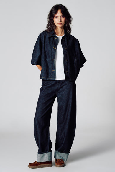 Manteau de denim bleu marine de Le Fabuleux Marcel de Bruxelles, porté sur un chemise blanche et un pantalon de denim large avec ceinture.