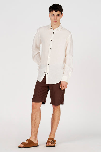 Chemise blanche en lin à manches longues de Le Fabuleux Marcel de Bruxelles, avec boutons foncés, combinée à un short marron.