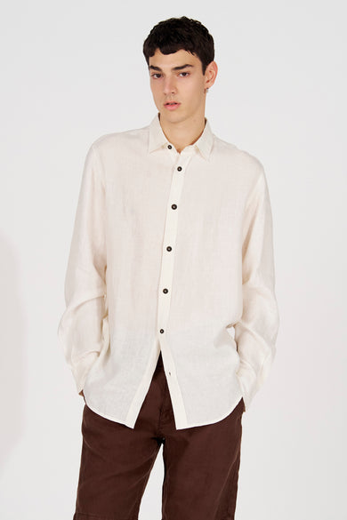Chemise en lin blanc à manches longues de Le Fabuleux Marcel de Bruxelles, non boutonnée, avec boutons foncés et pantalon marron.