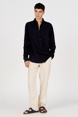 Chemise bleu foncé à manches longues de Le Fabuleux Marcel de Bruxelles, avec col Henley et boutons, portée avec un pantalon beige et des sandales.