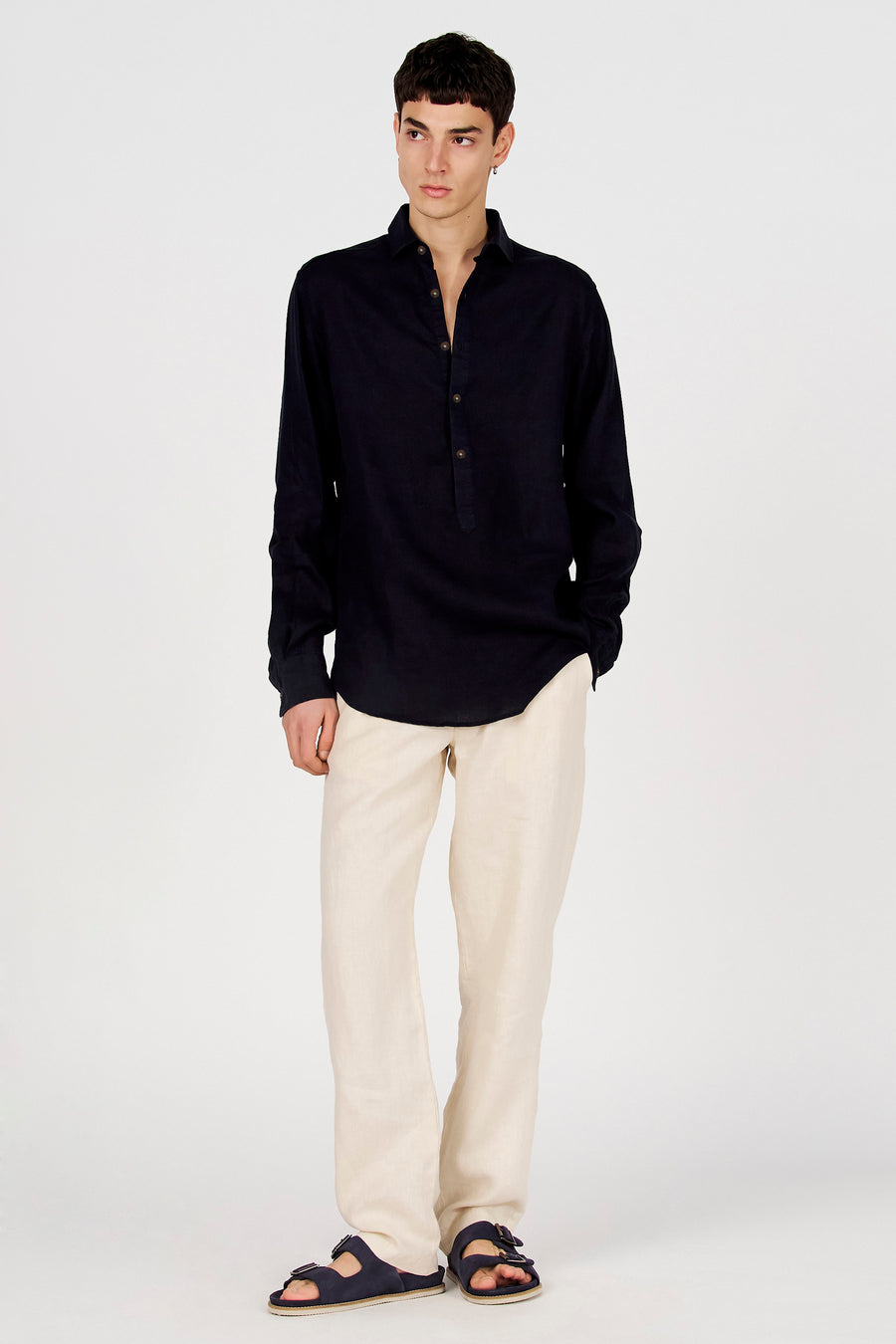 Chemise bleu foncé à manches longues de Le Fabuleux Marcel de Bruxelles, avec col Henley et boutons, portée avec un pantalon beige et des sandales.