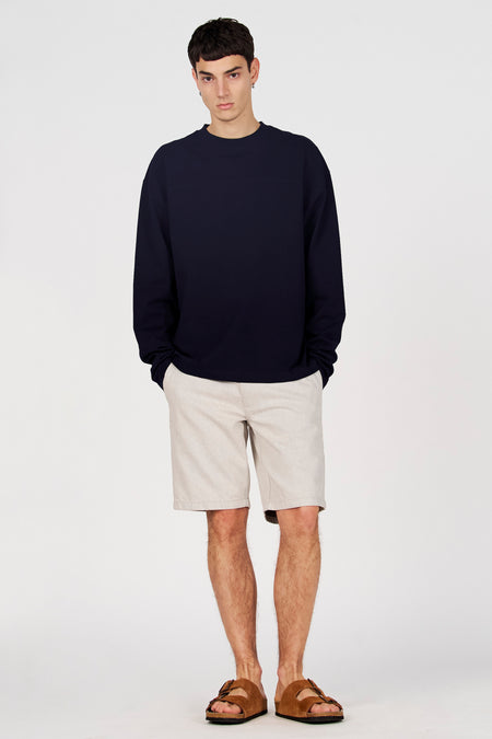 Pull bleu de Le Fabuleux Marcel de Bruxelles, porté avec un short beige et des sandales marron, debout devant un fond blanc.