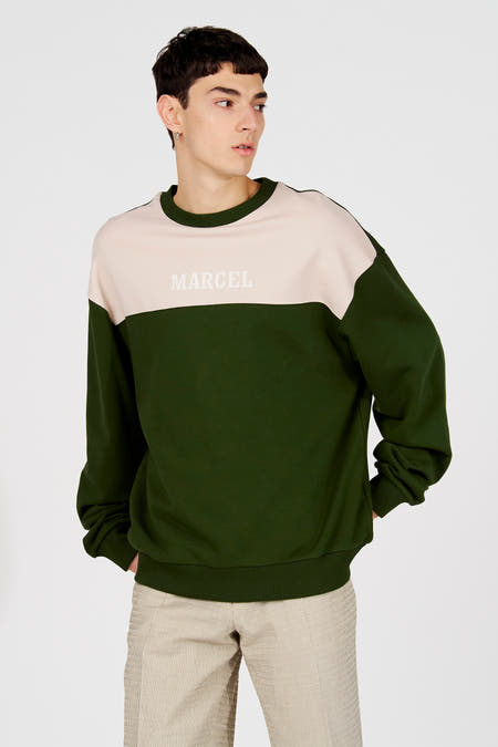 Groene sweater met ronde hals van Le Fabuleux Marcel de Bruxelles, met een lichtroze baan over de borst en het woord "MARCEL" erop.
