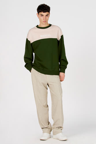 Pull vert à col rond de Le Fabuleux Marcel de Bruxelles, avec empiècement beige et le nom de la marque dessus, porté avec un pantalon rayé et des chaussures blanches.