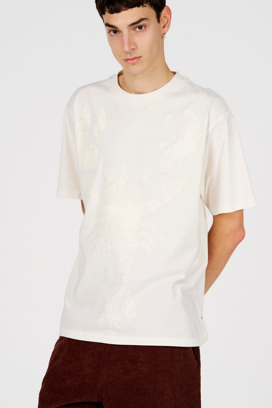 T-shirt blanc (manches courtes) de Le Fabuleux Marcel de Bruxelles, avec un motif graphique sur le devant, combiné avec un pantalon marron.