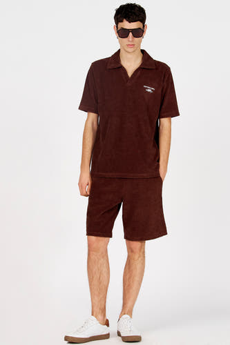 Polo marron de Le Fabuleux Marcel de Bruxelles à manches courtes avec un logo, portée avec un short assorti et des baskets blanches.