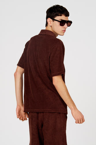 Bruine polo van Le Fabuleux Marcel de Bruxelles, met een subtiel textuurpatroon, gezien van achteren, gecombineerd met bijpassende broek en zwarte zonnebril.
