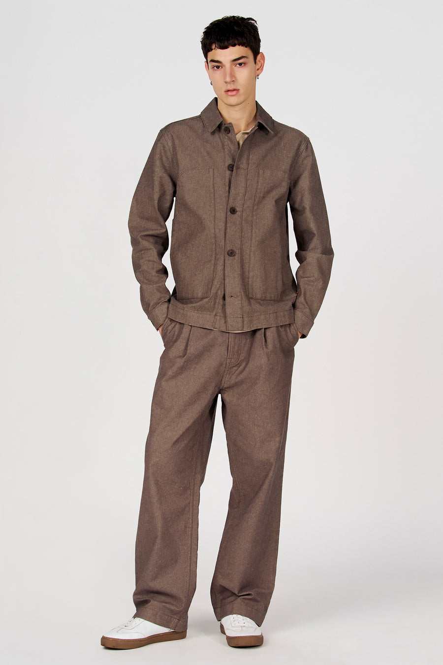 Veste courte marron de Le Fabuleux Marcel de Bruxelles, avec manches longues et boutons marron, portée avec un pantalon marron assorti et des chaussures blanches.