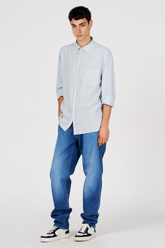 Chemise bleue à manches longues de Le Fabuleux Marcel de Bruxelles, avec poche poitrine et portée avec un jean bleu et des baskets.