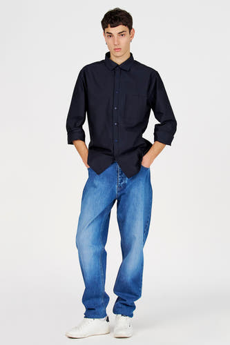Chemise bleue à manches longues de Le Fabuleux Marcel de Bruxelles, avec fermeture boutonnée et poche poitrine, portée avec un jean bleu et des baskets blanches.