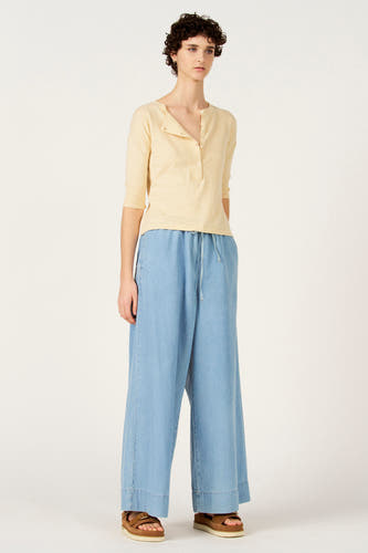 Pull écru à col rond de Le Fabuleux Marcel de Bruxelles, à manches courtes et fermeture boutonnée. Porté avec un pantalon ample bleu clair et des sandales.