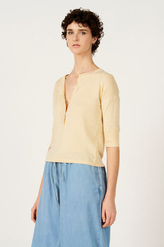 Pull écru à col rond de Le Fabuleux Marcel de Bruxelles, à manches courtes et porté avec un pantalon bleu à cordon de serrage.