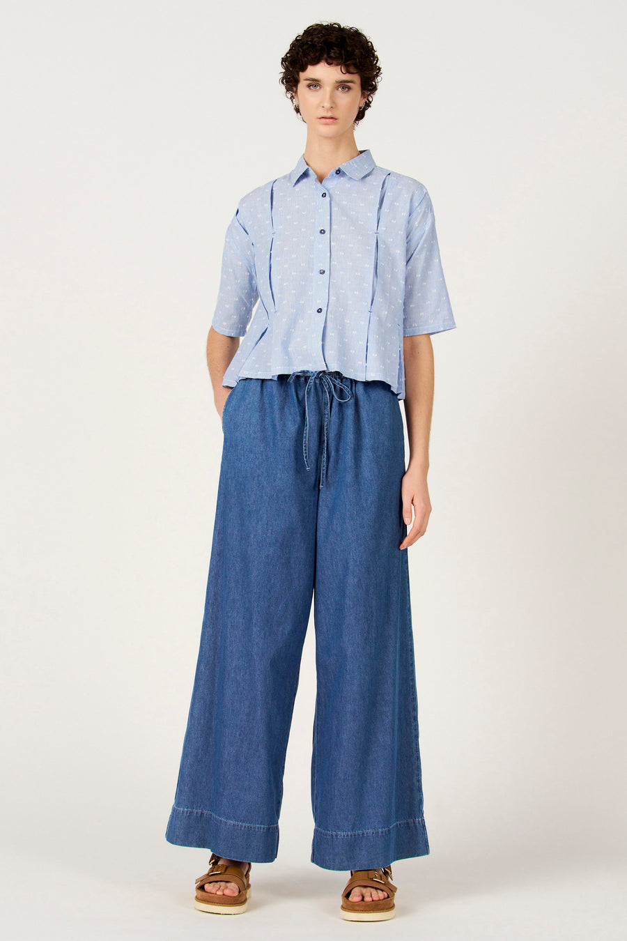 Chemise bleue à rayures à manches courtes de Le Fabuleux Marcel de Bruxelles, avec des pois blancs et portée avec un pantalon ample bleu foncé.