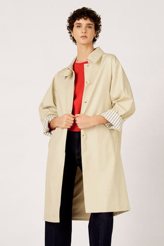 Long manteau beige de Le Fabuleux Marcel de Bruxelles, avec des manchettes rayées, porté sur un pull rouge et un jean foncé.