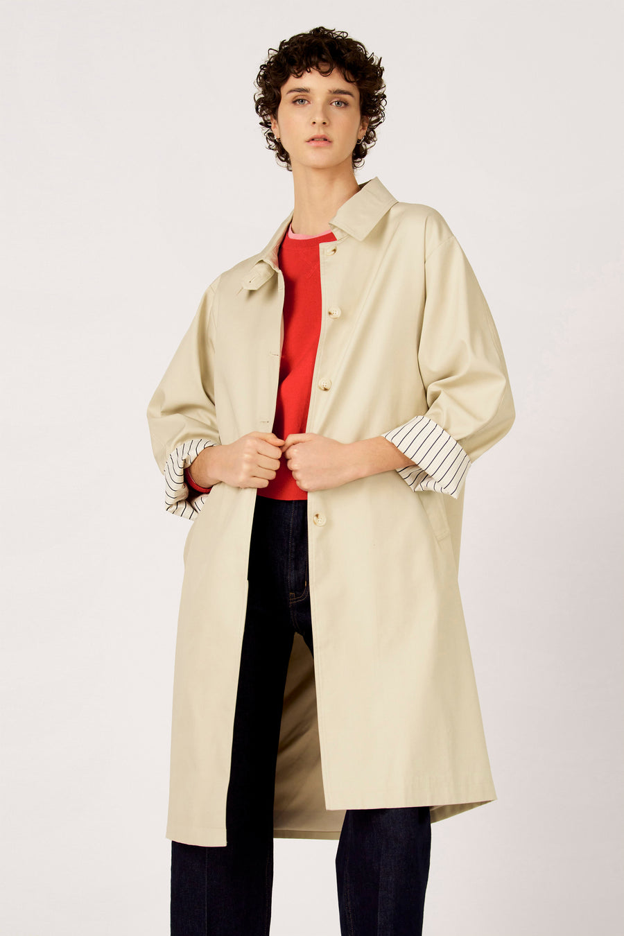 Long manteau beige de Le Fabuleux Marcel de Bruxelles, avec des manchettes rayées, porté sur un pull rouge et un jean foncé.