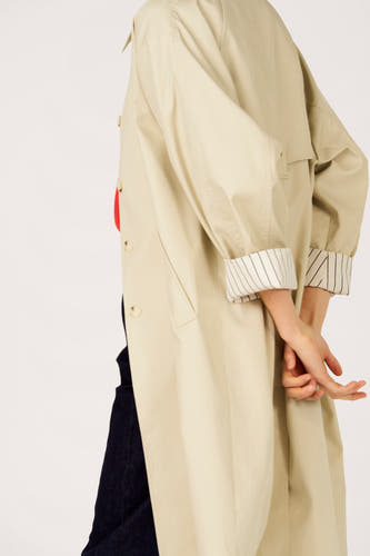 Long manteau beige de Le Fabuleux Marcel de Bruxelles, avec revers rayé, haut rouge et pantalon foncé.